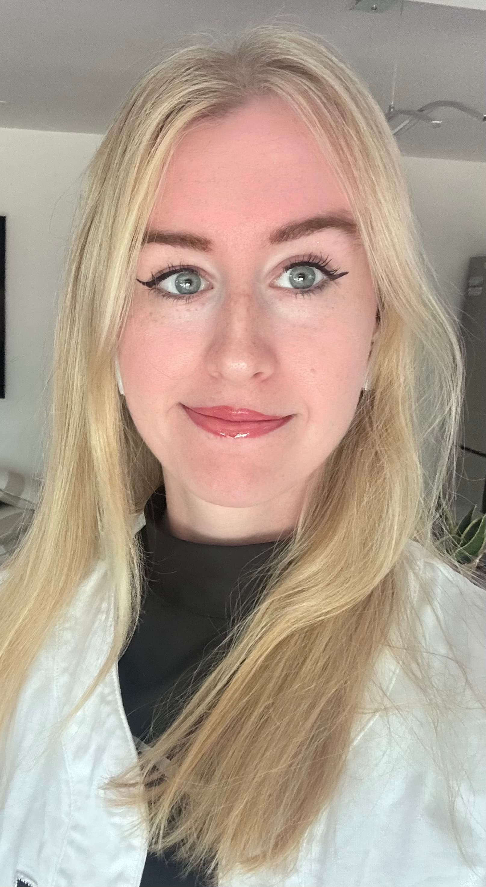 A blonde woman with blue eyes and winged eyeliner smiles softly in a close-up.